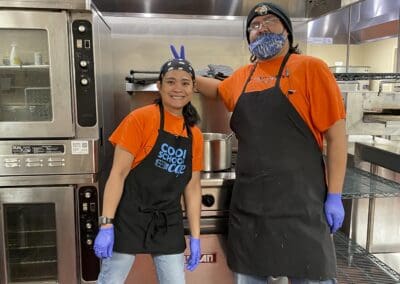 two cooks in the kitchen wearing orange shirts and black aprons.